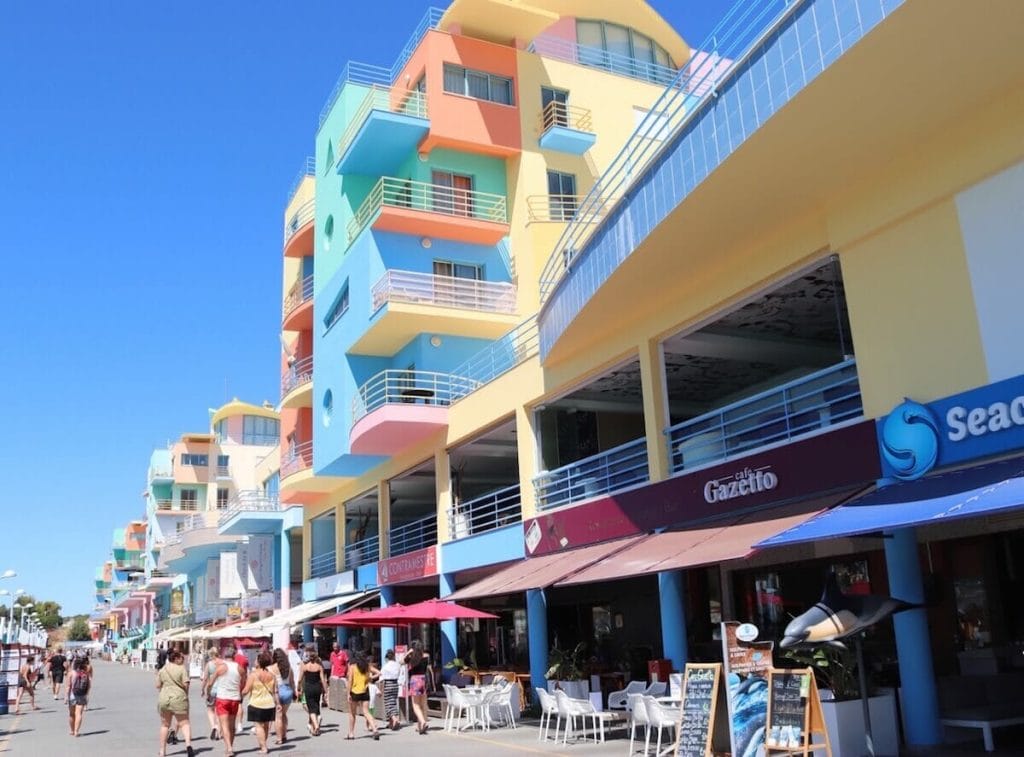 The Marina de Albufeira with its bars, restaurants with outside seatings, seven-storey colourful buildings, and some people walking