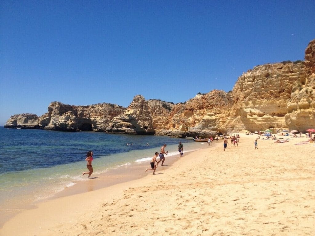 Pessoas caminhando na areia fofa de areia âmbar da Praia da Marinha, que é orlada por enormes falésias amarelo-laranjas e tem águas cristalinas de um azul. 