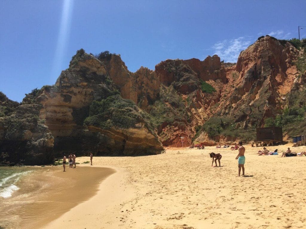 enormes falésias vermelhas da Praia do Camilo e algumas pessoas de pé na areia cor de âmbar