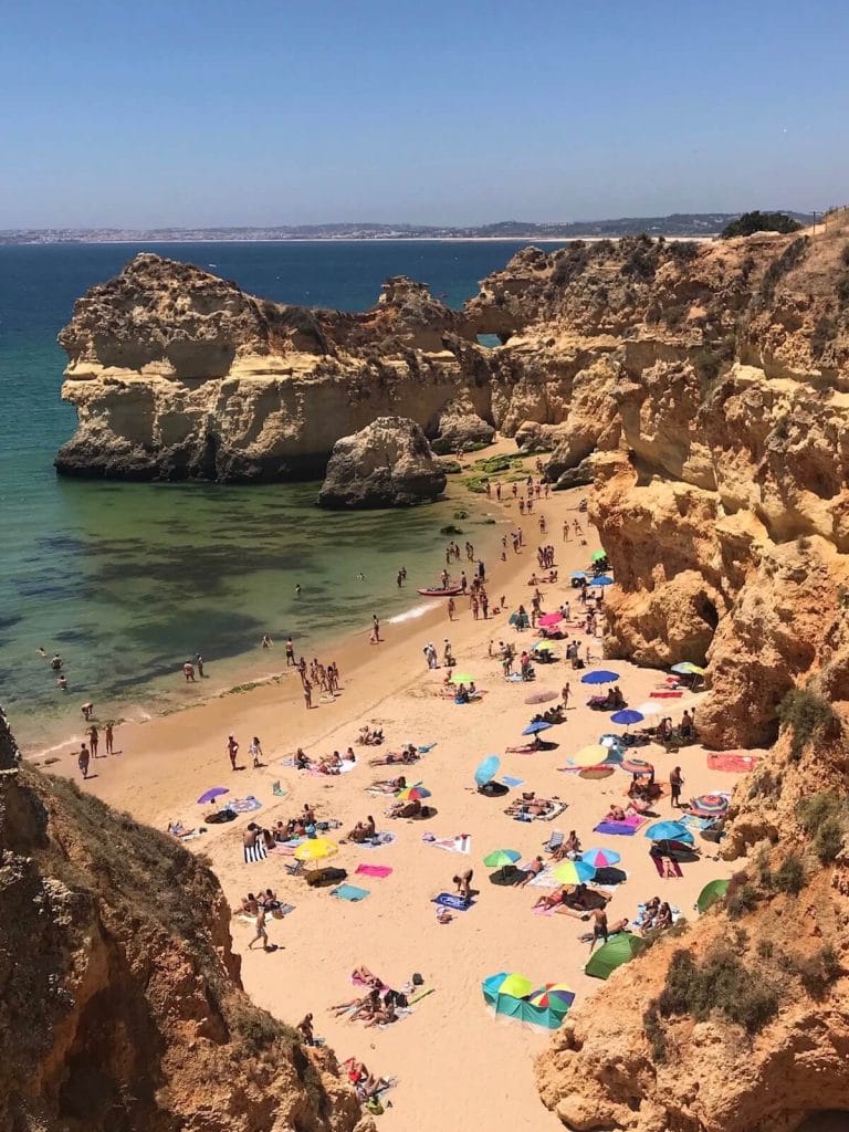 Algunas personas caminando, nadando y tomando el sol bajo sombrillas de diferentes colores en la Praia dos Três Irmãos, Portimão, que está rodeada de enormes acantilados de piedra caliza de color naranja amarillento y tiene agua verde cristalina.