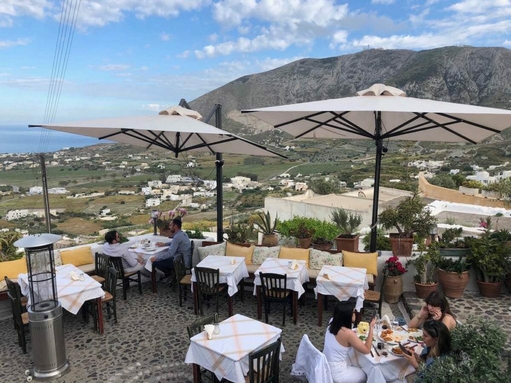 five people eating on the outside seating of Metaxi Max Restaurant, Santorini, which has white umbrellas, tables, chairs and some plants 