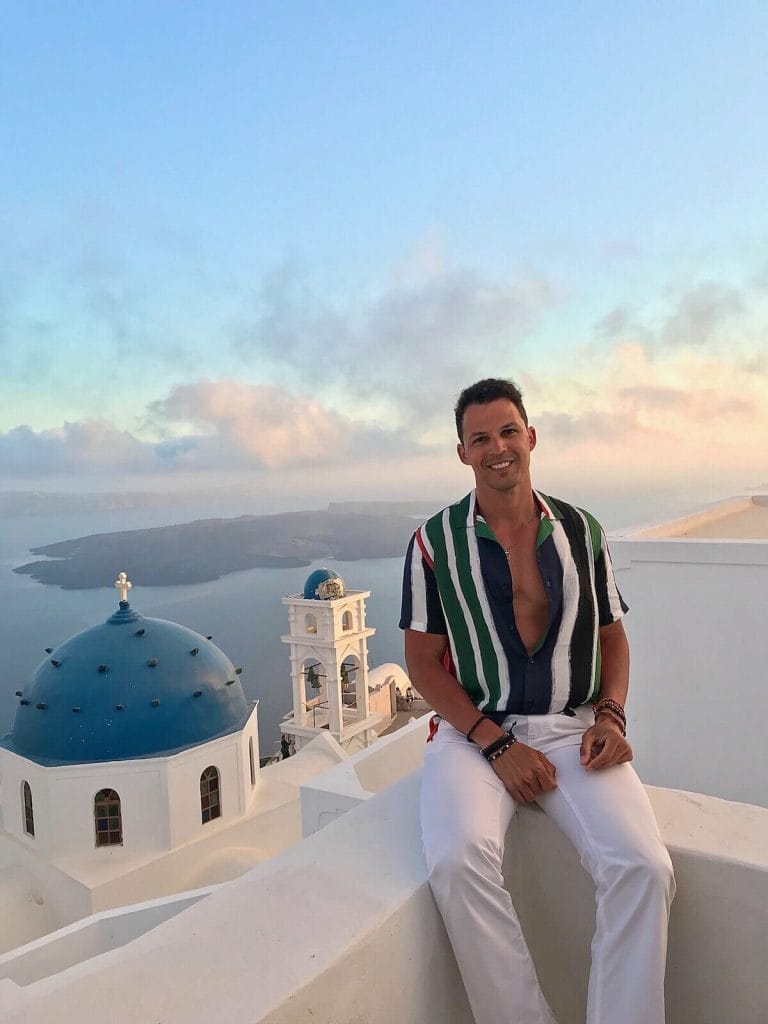 Pericles Rosa wearing a striped shirt and white trouser sitting on a white balcony in the village of Imerovigli, Santorini, and ti the background the Anastasi Church, Nea Kammeni island and the Aegean Sea