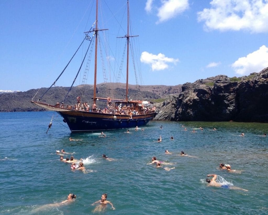 Un barco de madera lleno de gente cerca de la isla Nea Kameni, en Santorini, y algunas personas nadando en el Mar Egeo