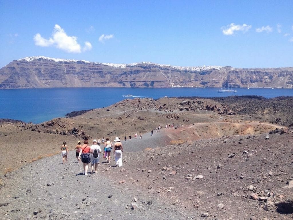 people walking at Nea Kameni Volcanic Park, which has black sand and volcanic rocks, and the island of Santorini with whitewashed houses atop of an immense cliff in the background