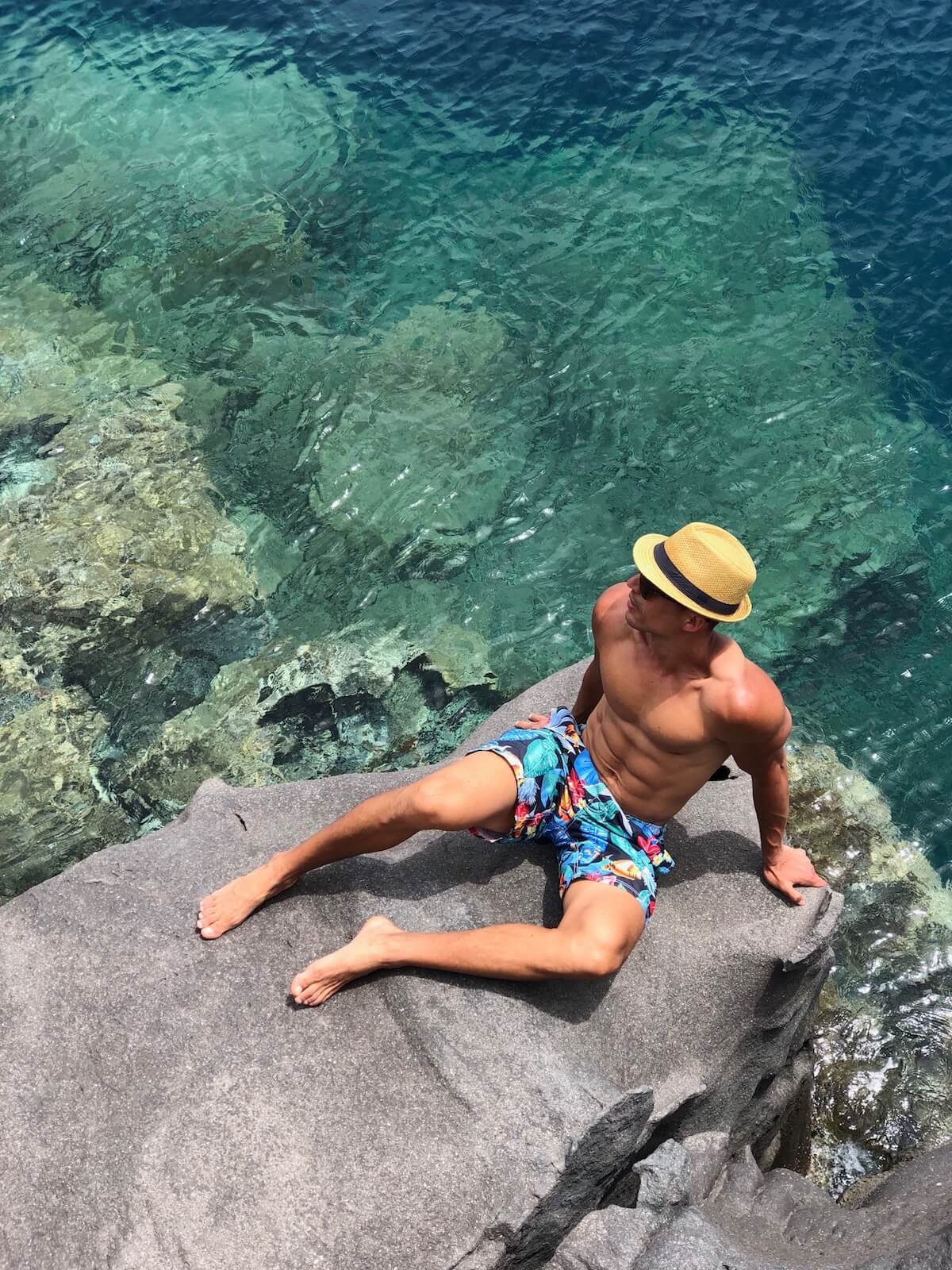Un hombre usando un sombrero, gafas de sol y pantalones cortos coloridos sentado en una roca en la Bahía de Ammoundi, Santorini, Grécia