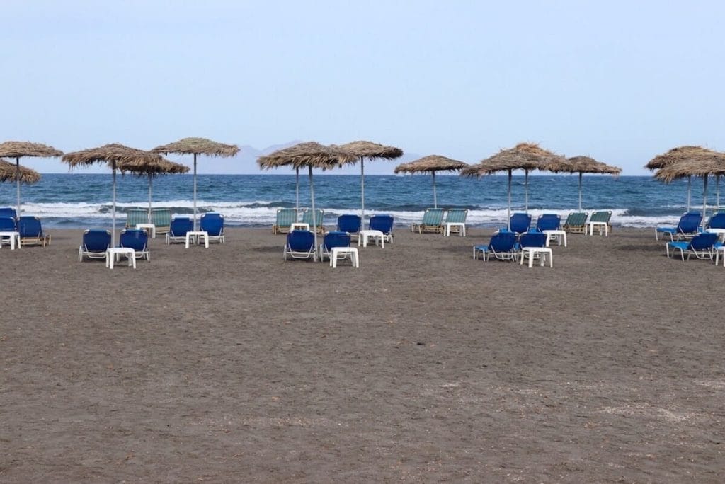 A stretch of black sand with umbrellas and beaches chairs on Monolithos Beach, Santorini, Greece