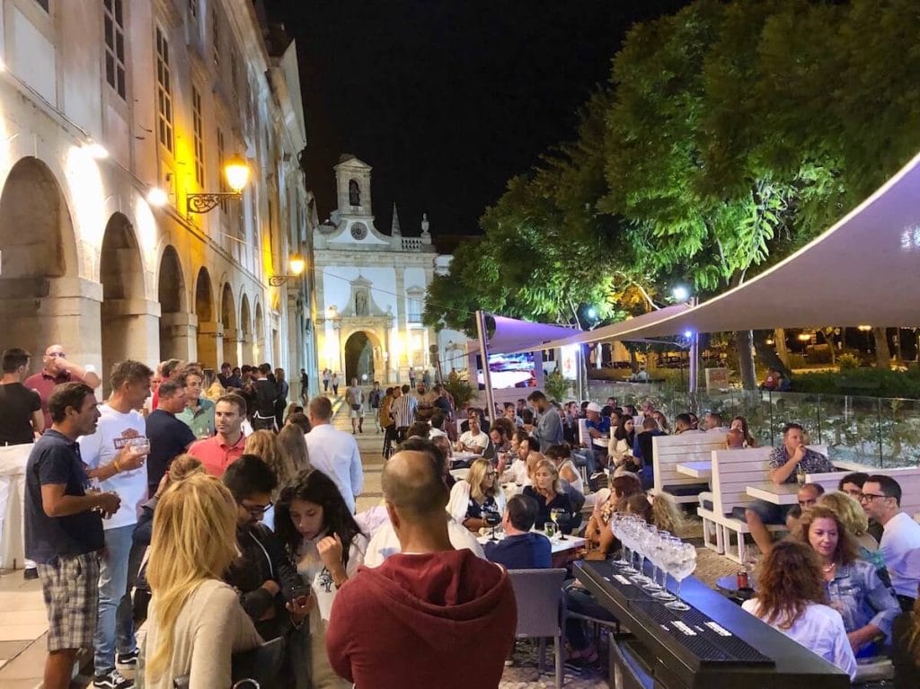 Pessoas sentadas em cadeiras e outras de pé no Bar Columbus no centro histórico de Faro, Portugal