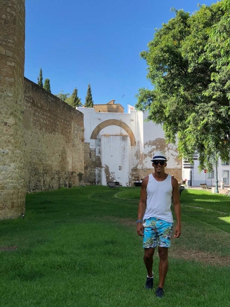 un hombre con un sombrero blanco, una camiseta sin mangas blanca y pantalones cortos de colores caminando cerca de las antiguas murallas del casco antiguo de Faro