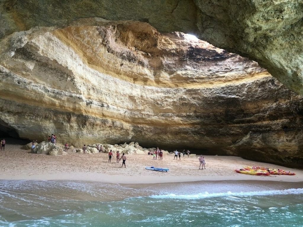 Some kayaks and people walking inside Benagil Cave, Portugal, that has golden-cloured walls and a round hole in the ceiling