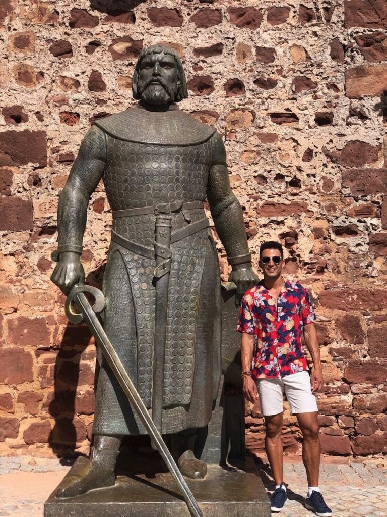 Pericles Rosa vistiendo pantalones cortos beige y una camisa de flores roja con una enorme estatua del Rey Sancho I en la entrada del Castelo de Silves, Portugal