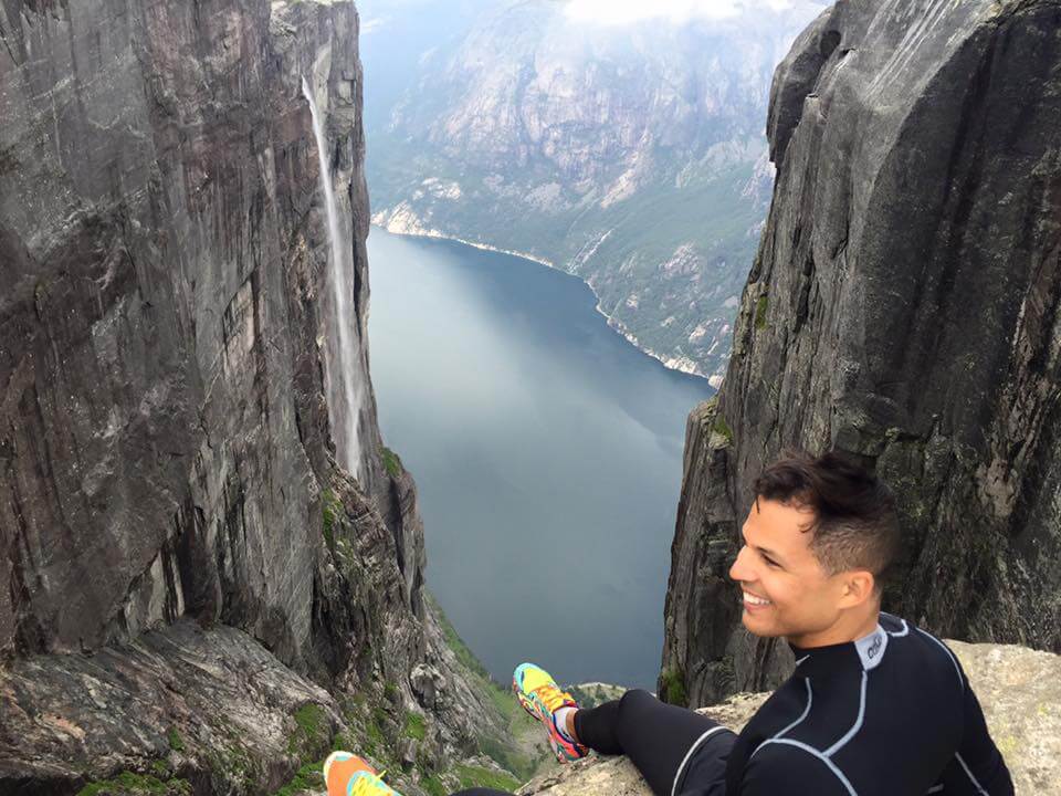 Pericles Rosa wearing black clothes and colourful trainers sitting on the edge on the top of Kjerag Mountain admiring the view of the Lysefjorden fjord and the Kjeragfossen waterfall that plunges off the mountain down to the fjord