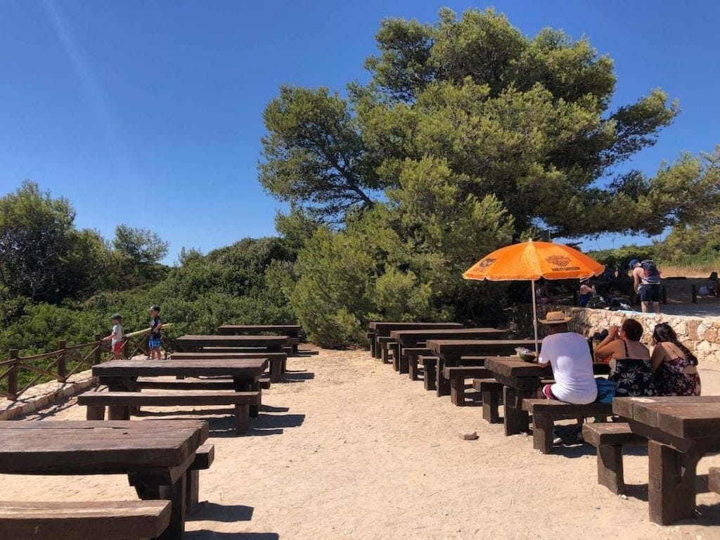 A picnic area at the end of The Seven Hanging Valleys Trail in the Algarve, Portugal, with wooden tables and benches, some trees and people in one of the tables with an orange umbrella drinking and eating