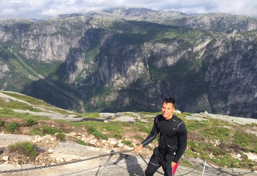 Pericles Rosa wearing black clothes and carrying a black backpack holding onto some chains during while hiking up to Kjeragbolten