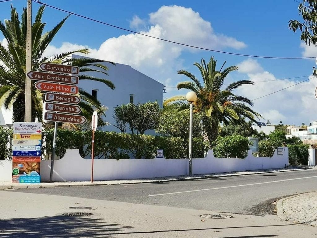 Some signs on the corner of a street showing how to get to Marinha Beach, Carvoeiro, Benagil and Albandeira, a white house and trees in the background