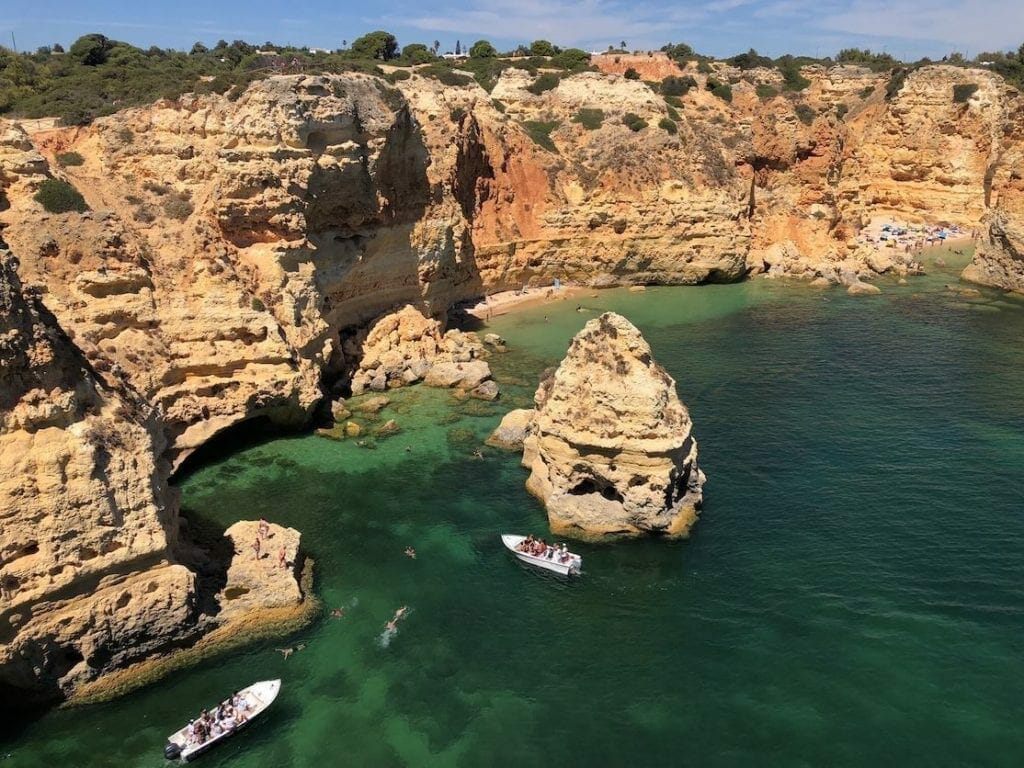 The extraordinary Marinha Beach, one of the most beautiful beaches in the Algarve, Portugal, with people swimming and two boats on its crystalline blue water bordered by orange-yellowish cliffs