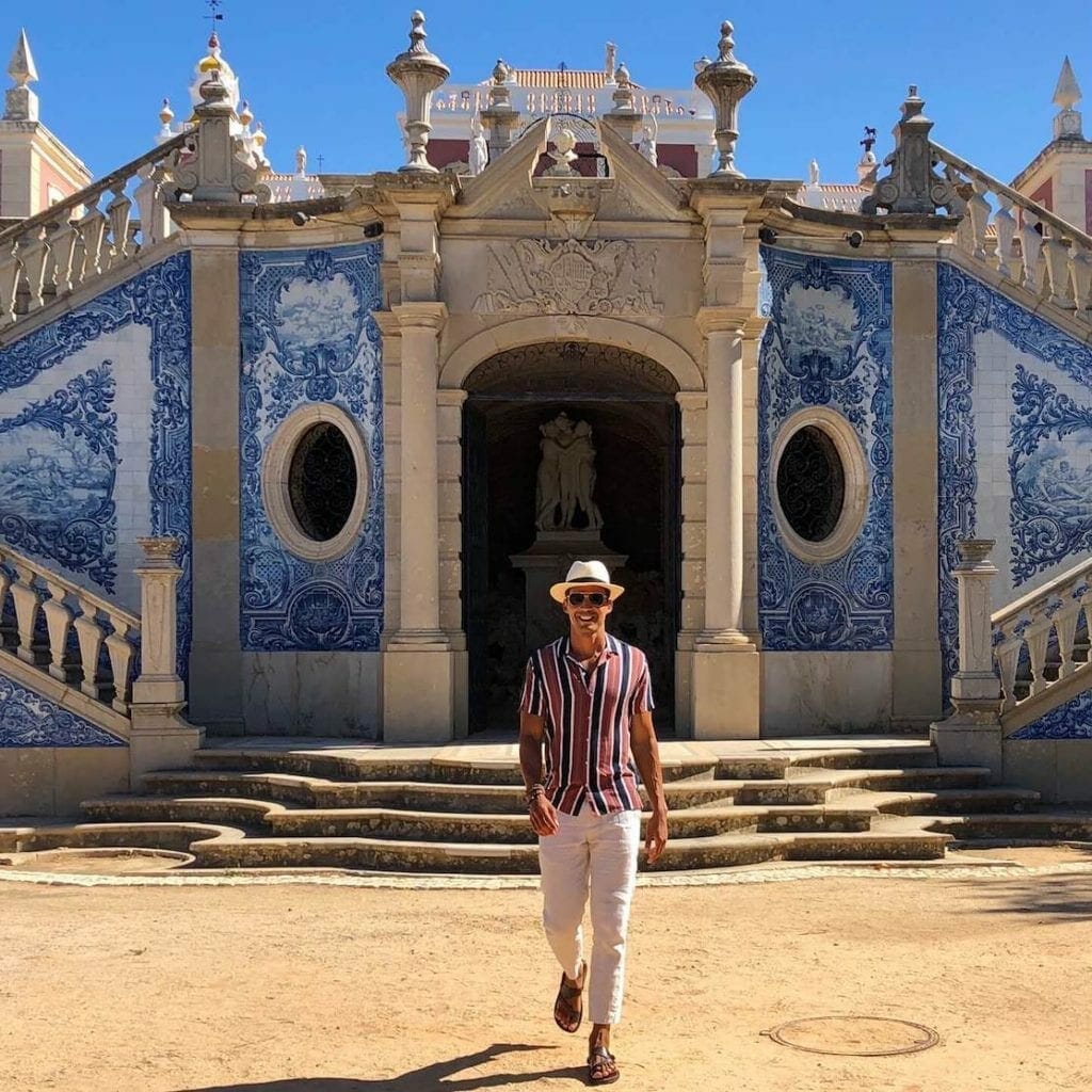 A man wearing beige trousers, striped brown shirt, sunglasses and beige hat walking at Palacio de Estói, in Faro, with a wall covered in Portuguese blue tiles behind him