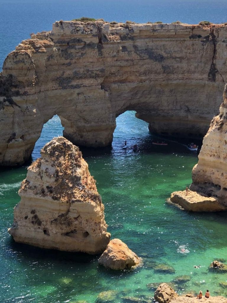 Una playa de agua esmeralda cristalina con enormes acantilados de piedra caliza amarilla, incluido uno con dos arcos marinos, gente haciendo Stand-up Paddle Boarding y una pareja sentada en uno de los acantilados admirando la vista