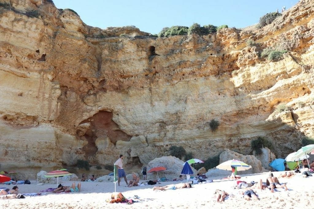 People sunbathing on towels and underneath umbrellas on Praia da Marinha with an immense yellow limestone cliff in the background