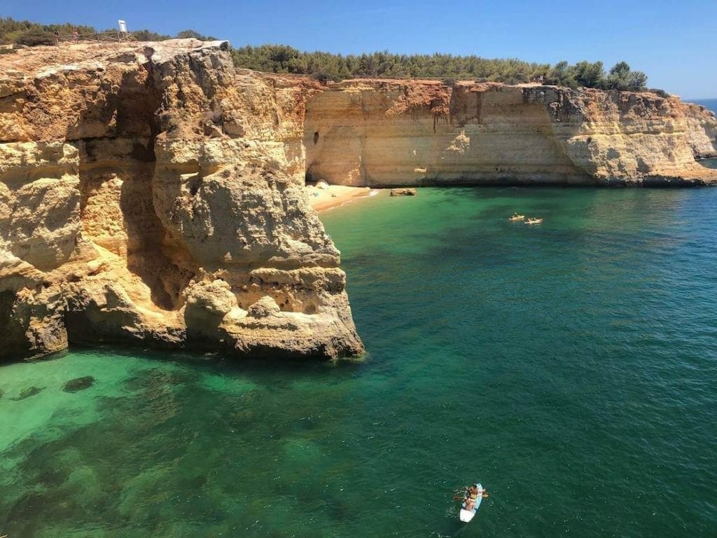 A beach in the Algarve, Portugal, surrounded by yellow cliffs covered with low vegetation, and three kayaks sailing on its crystalline emerald water