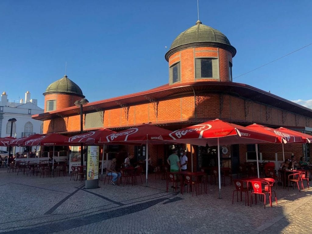 Olhão Market faced in red brick with some red plastic tables, chairs and umbrellas in front of it