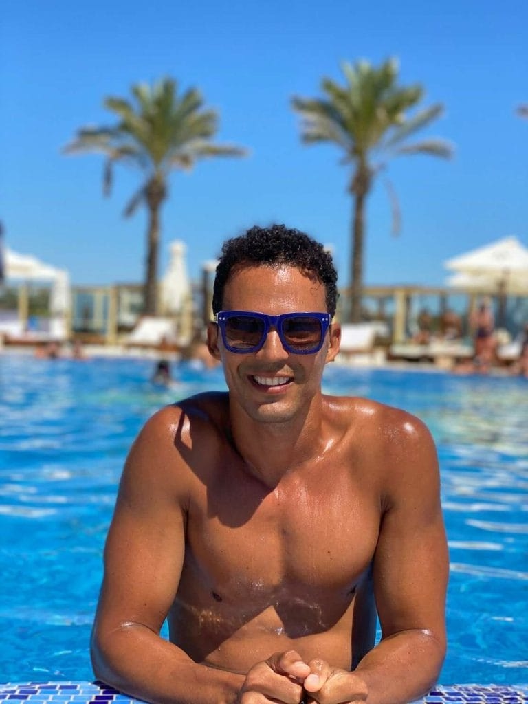 Pericles Rosa wearing blue sunglasses standing in the swimming pool of No Solo Agua Beach Club in Portimão, with two palm trees, some people, lounge chairs and white umbrellas in the background