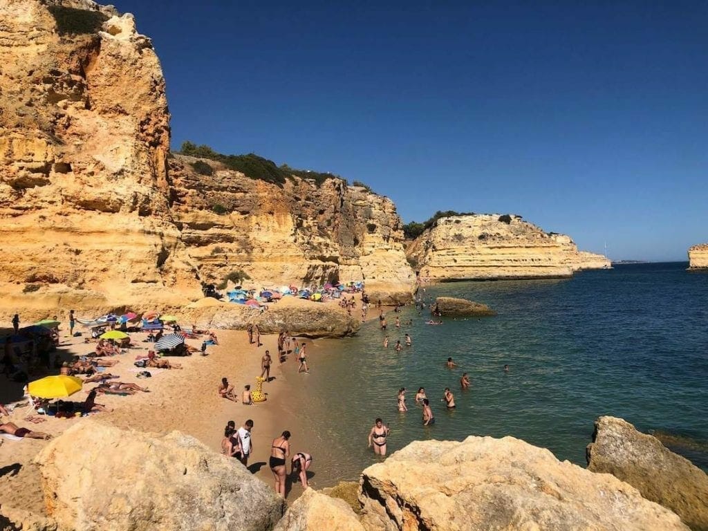 A beach bordered by cliffs and some people sunbathing, others walking on the beach and some more in the water