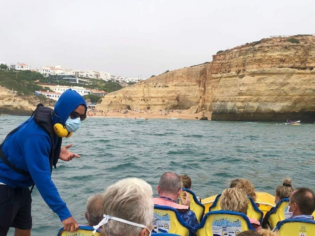 A tour guide wearing a blue hoodie jacket, sunglasses and a face mask giving some explanation to tourists sitting on yellow seats on a boat during a Dolphin Watching and Caves tours in the Algarve, Portugal