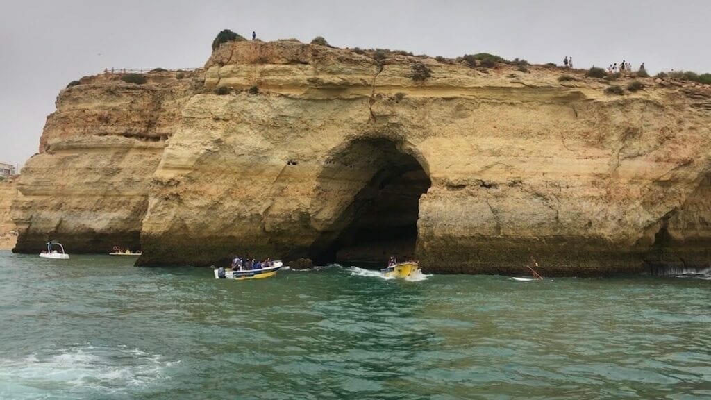 Four boats sailing around Algar de Benagil, Portugal, that has massive yellow limestone cloffs and some people walking on the top of the cliffs