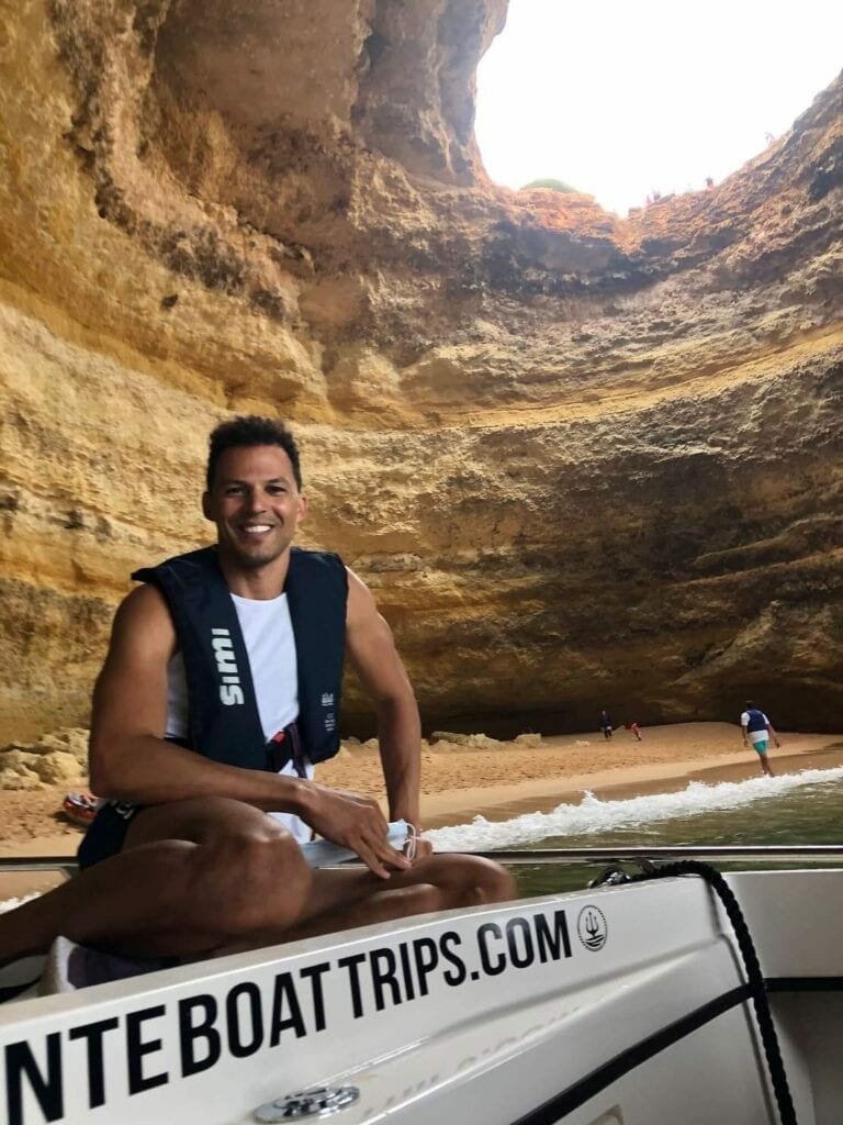 Pericles rosa wearing a white tank top and a black lifejacket sitting on a boat inside Benagil Cave, Portugal, and the cave's wall and round hole in its ceiling in the background