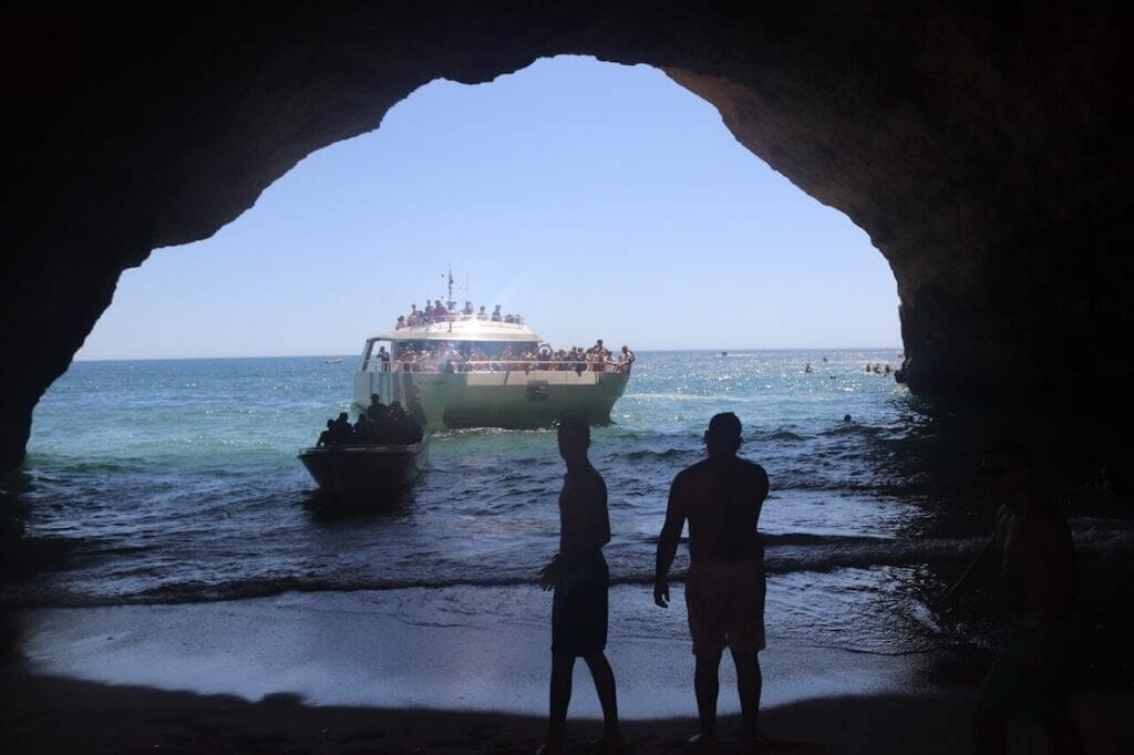 Two men inside of Benagil Cave and a catamaran and a small boat at its entrance 