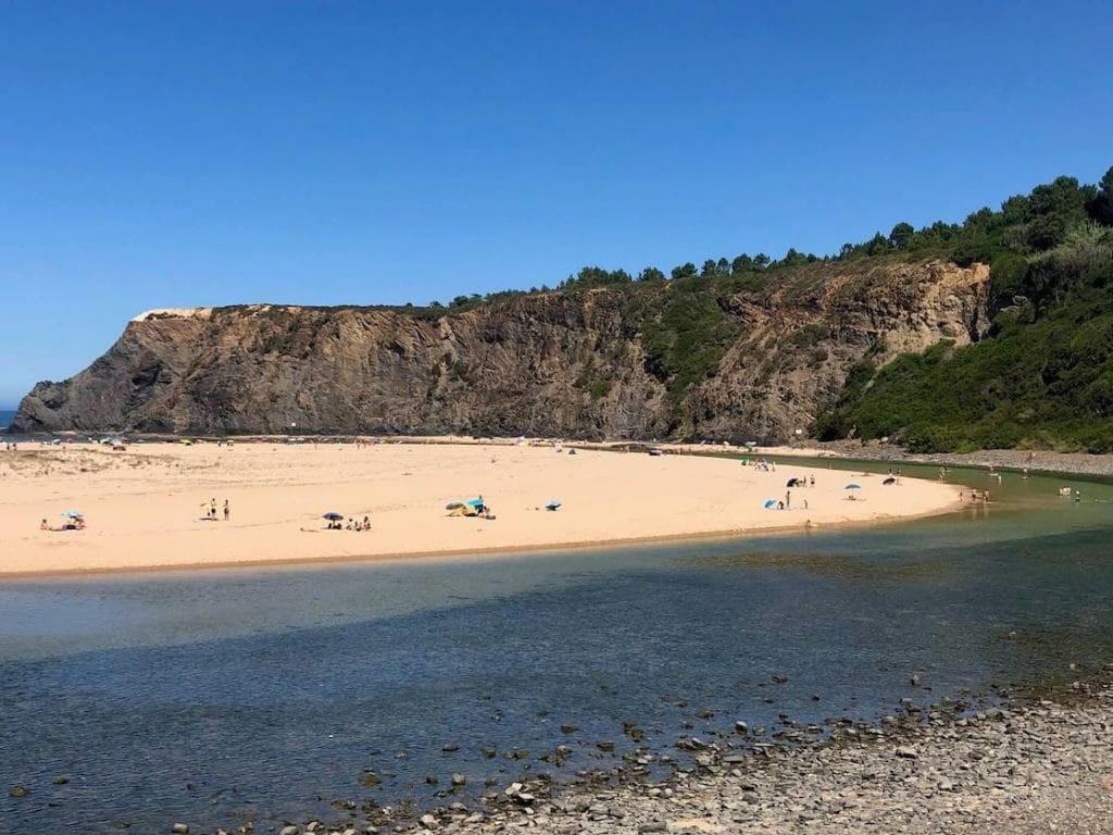 La playa de Odeceixe, Portugal, y su tramo de arena de forma redonda, un río de agua cristalina que la rodea y el acantilado negro cubierto de vegetación que bordea la playa