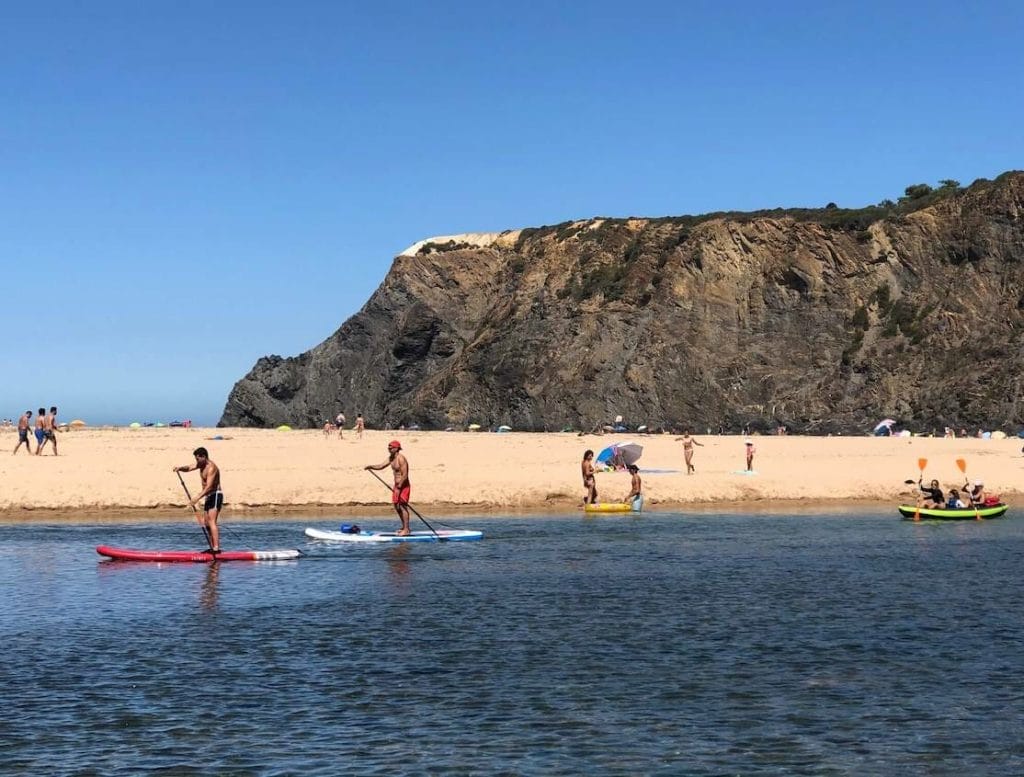 Pessoas praticando SUP e numa boia insuflável remando ao longo de um rio na Praia de Odeceixe, e outras caminhando na banco de areia e uma enorme falésia ao fundo