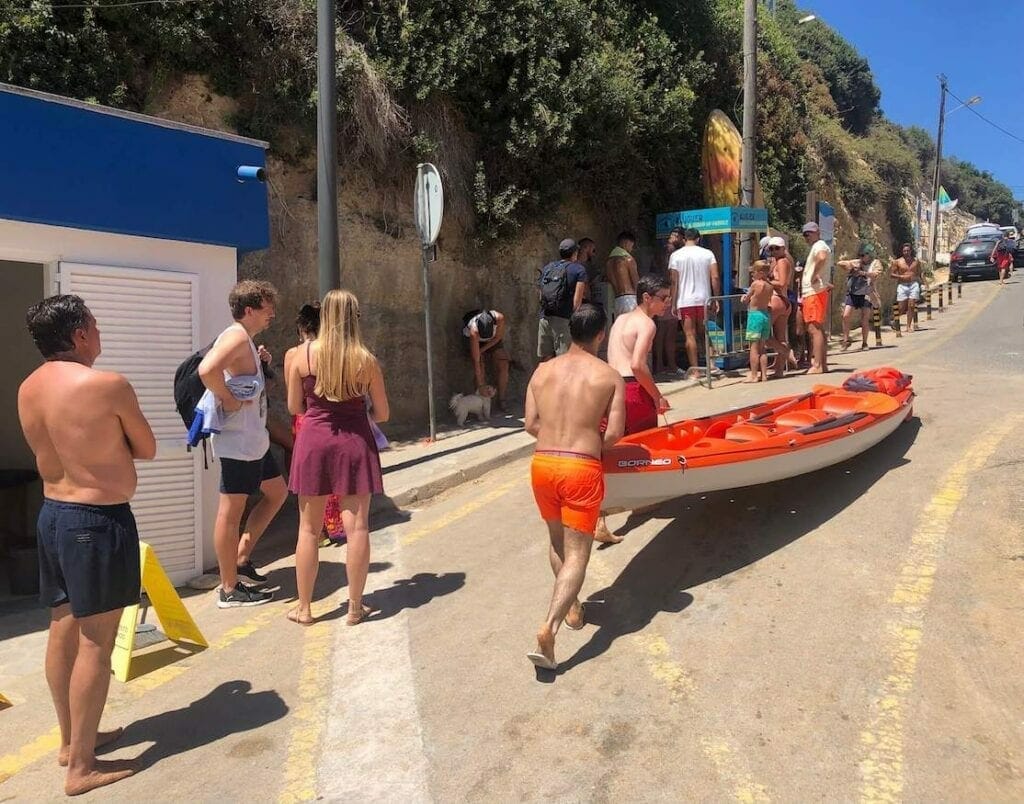 People waiting in line and two guys pushing a kayak on a street of Benagil, Portugal