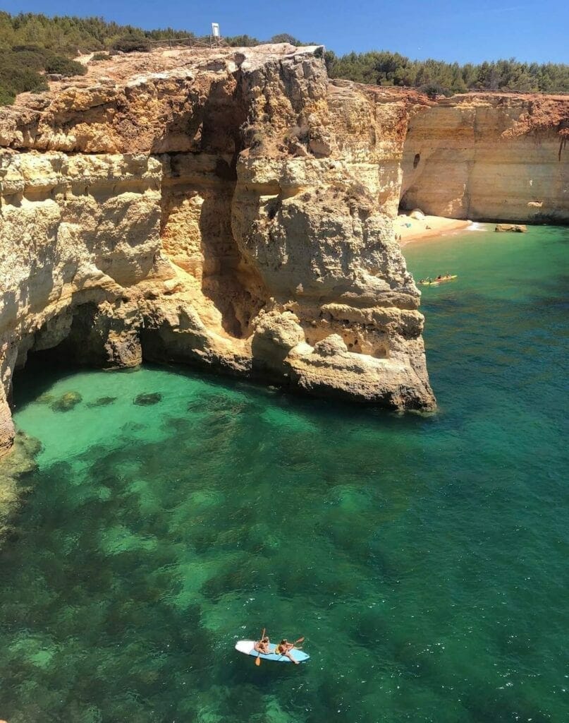 Two ladies on a kayak in the crystal-clear emerald water of Praia da Corredoura, Portugal, which is enveloped by tall yellow-orangeish limestone cliffs