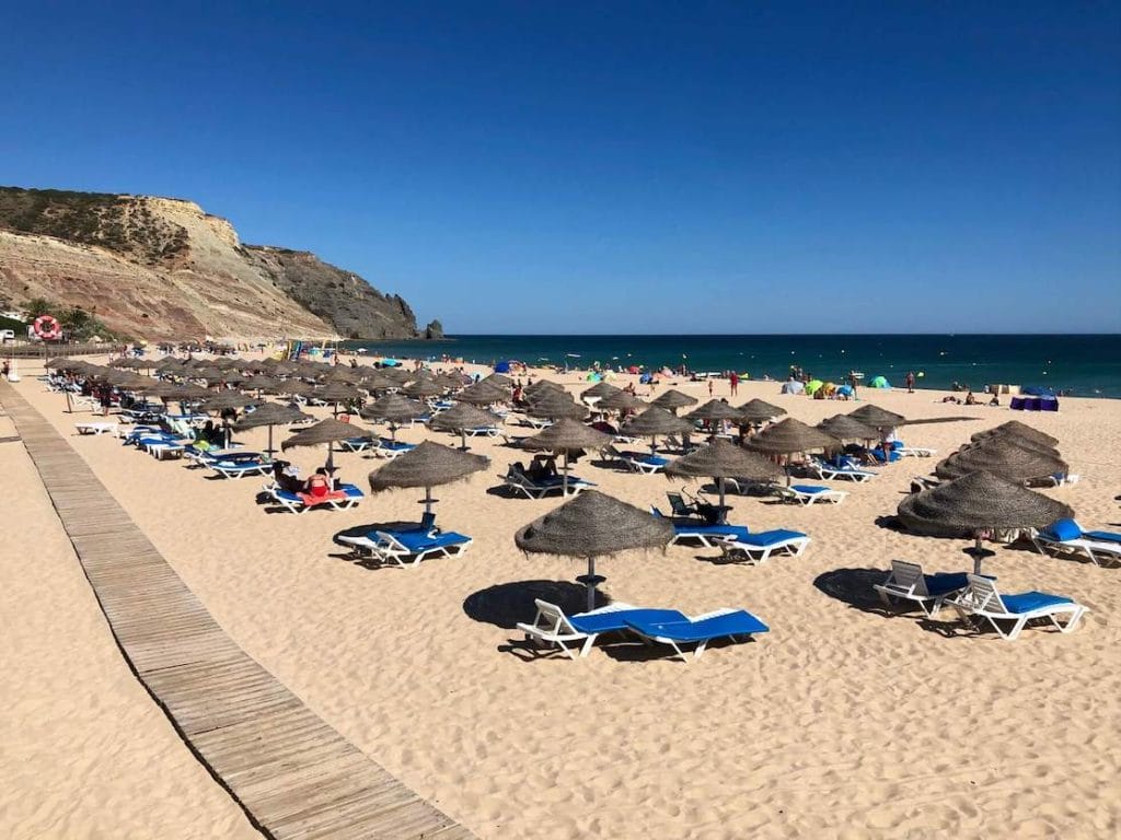 Um calçadão de madeira, guarda-sóis e espreguiçadeiras azuis na Praia da Luz, que tem areia fofa, águas cristalinas e é ladeada por uma imponente montanha