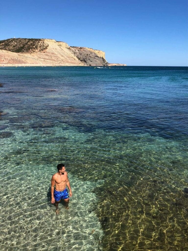 Un hombre vistiendo un pantalón corto de pie en el agua cristalina de Praia da Luz, Portugal, y el acantilado gris que bordea la playa al fondo