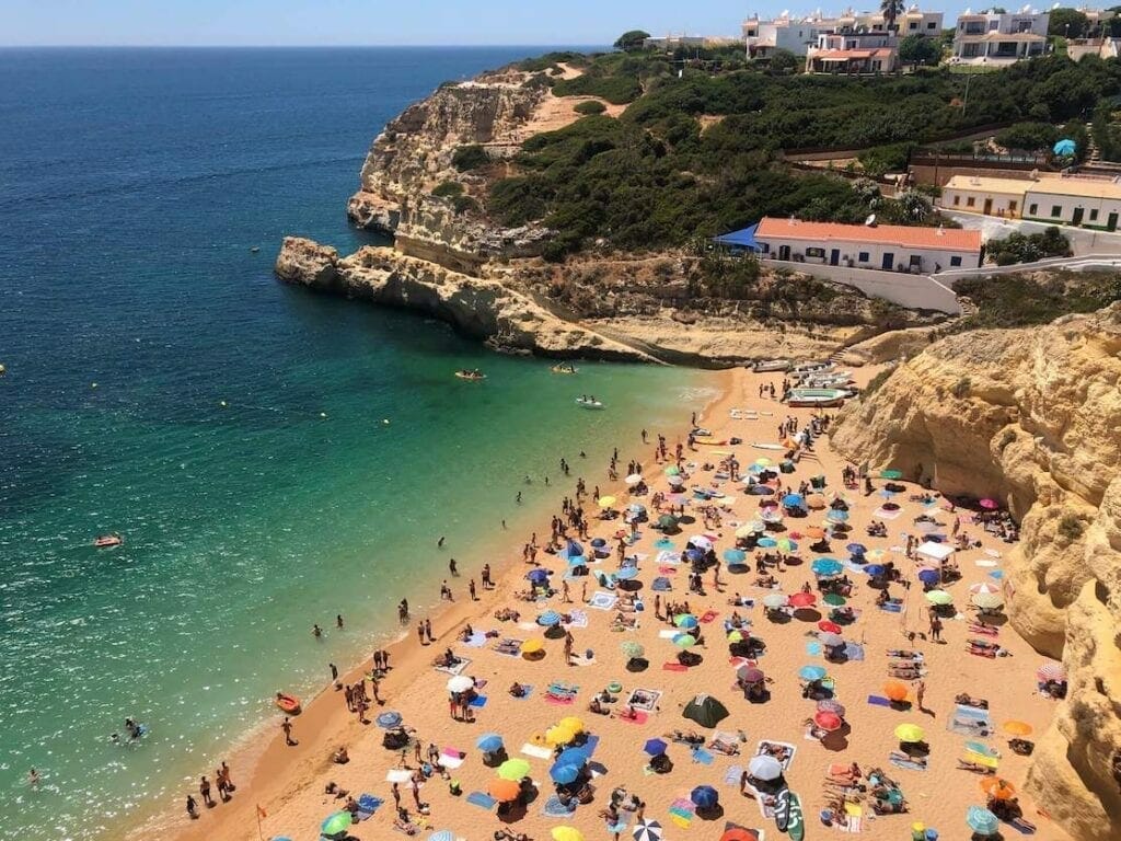 People sunbathing on the  towels and underneath umbrellas of different colours on Benagil Beach, Portugal, which is enveloped by orange-yellowish limestone cliffs and has crystal-clear blue water