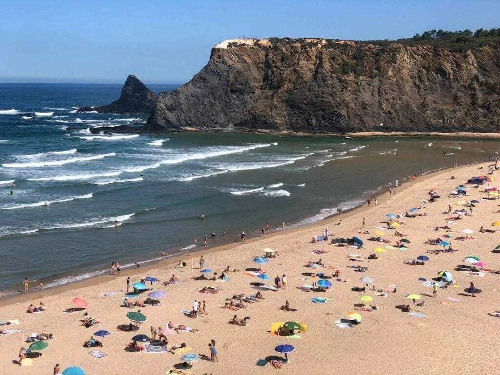 Personas tomando el sol en toallas y debajo de sombrillas de diferentes colores en la playa de Odeceixe, Portugal, el mar y un acantilado negro coronado con vegetación en el fondo