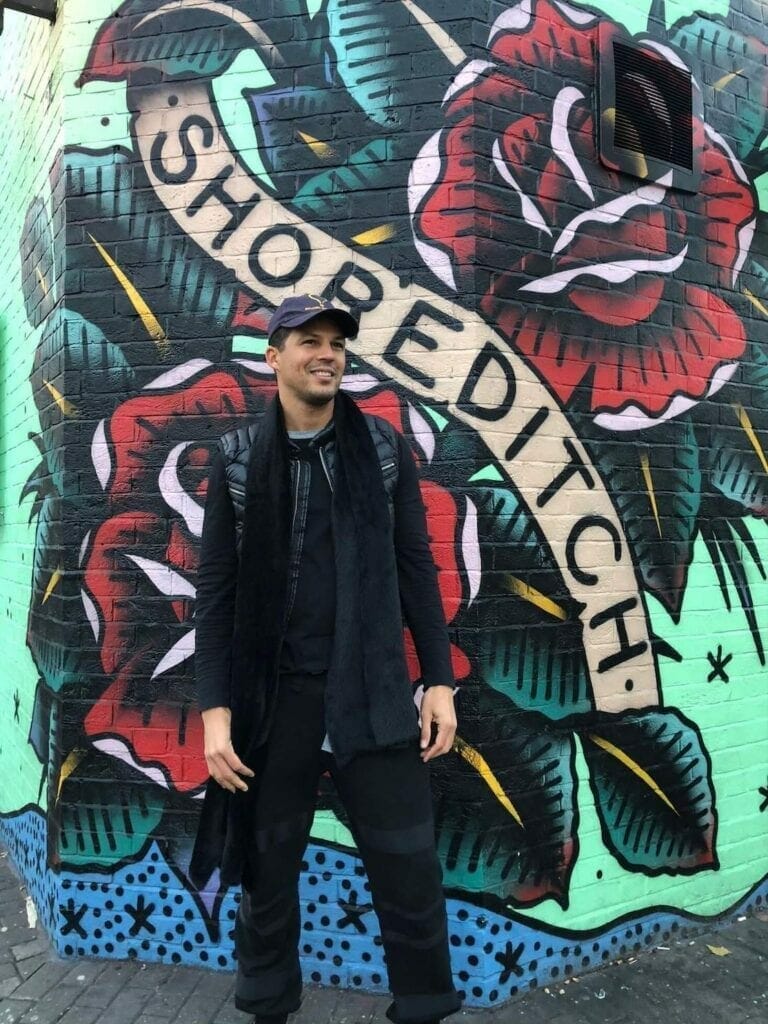 Um homem usando roupas pretas proximo a um mural de grafite no bairro de Shoreditch, Londres