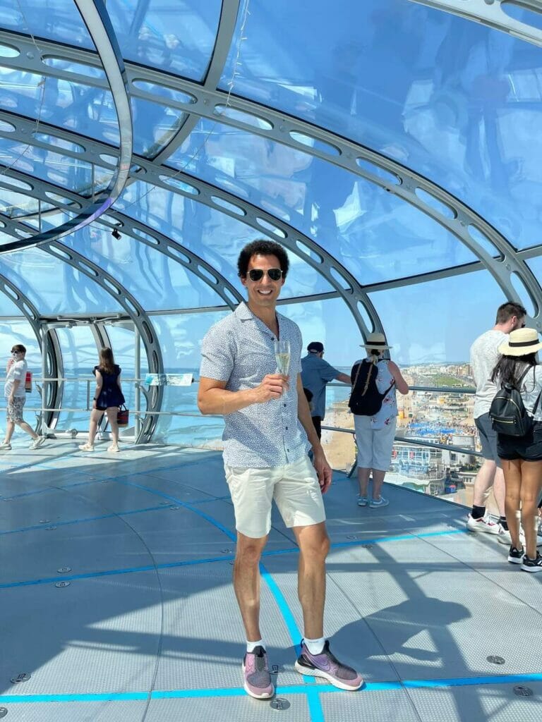 Pericles Rosa wearing sunglasses, a white shirt with blue dots, beige shorts and a pair of sneakers having a glass of sparkling wine inside the viewing pod of British Airways i360