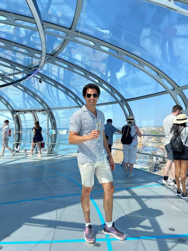 Péricles Rosa usando óculos escuros, uma camisa branca com pontos azuis, shorts bege e um par de tênis com uma taça de espumante dentro da cápsula de visualização da British Airways i360