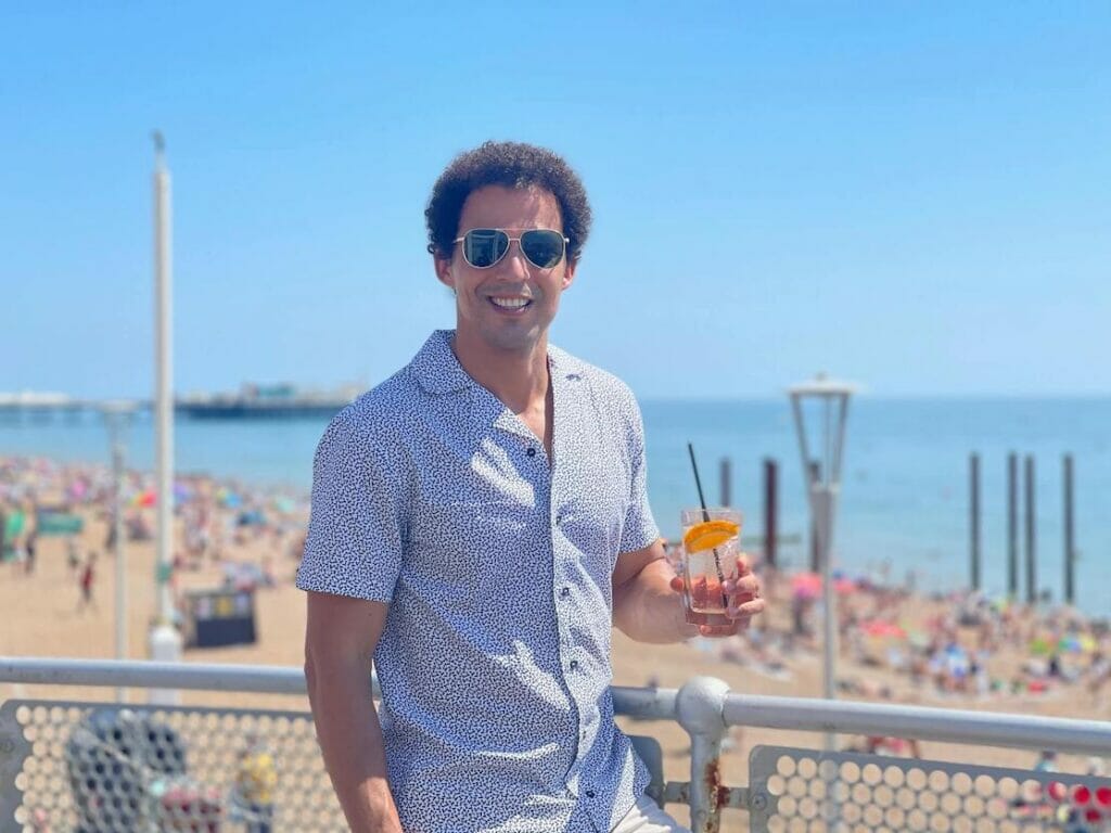 Pericles Rosa wearing sunglasses and white shorts dotted with blue dots having a drink at Brighton Beach Club and the beach behind him