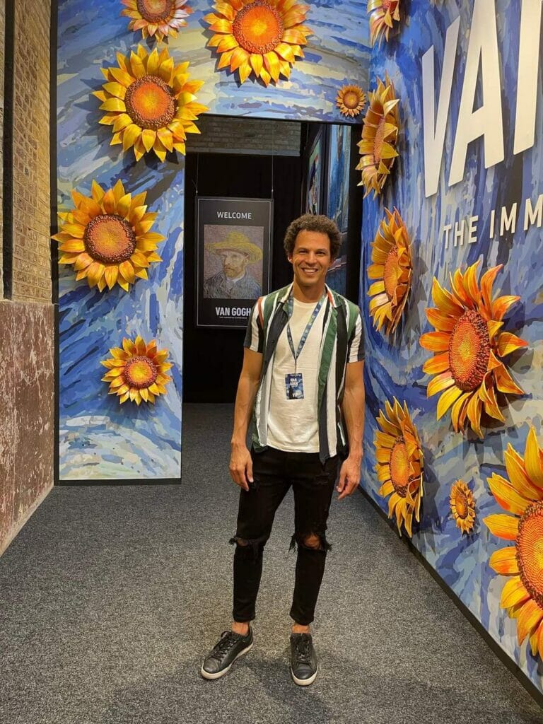 Pericles Rosa wearing black trousers and sneakers, white t-shirt and striped shirt standing at a hall with blue walls decorated huge sunflowers at Van Gogh exhibition, London