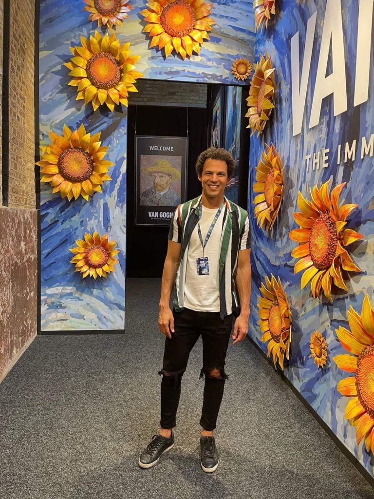 Péricles Rosa vestindo calças pretas e tênis, camiseta branca e camisa listrada em pé em um salão com paredes azuis decoradas enormes girassóis na exposição do Van Gogh, Londres