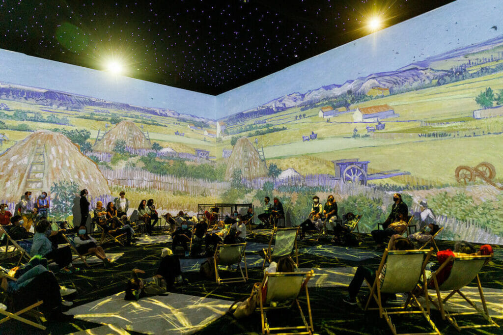 Uma sala com pessoas sentadas no chão e cadeiras de praia com paredes cobertas com paisagem bucólicas coloridas durante a Van Gogh: Experiência Imersiva em Londres