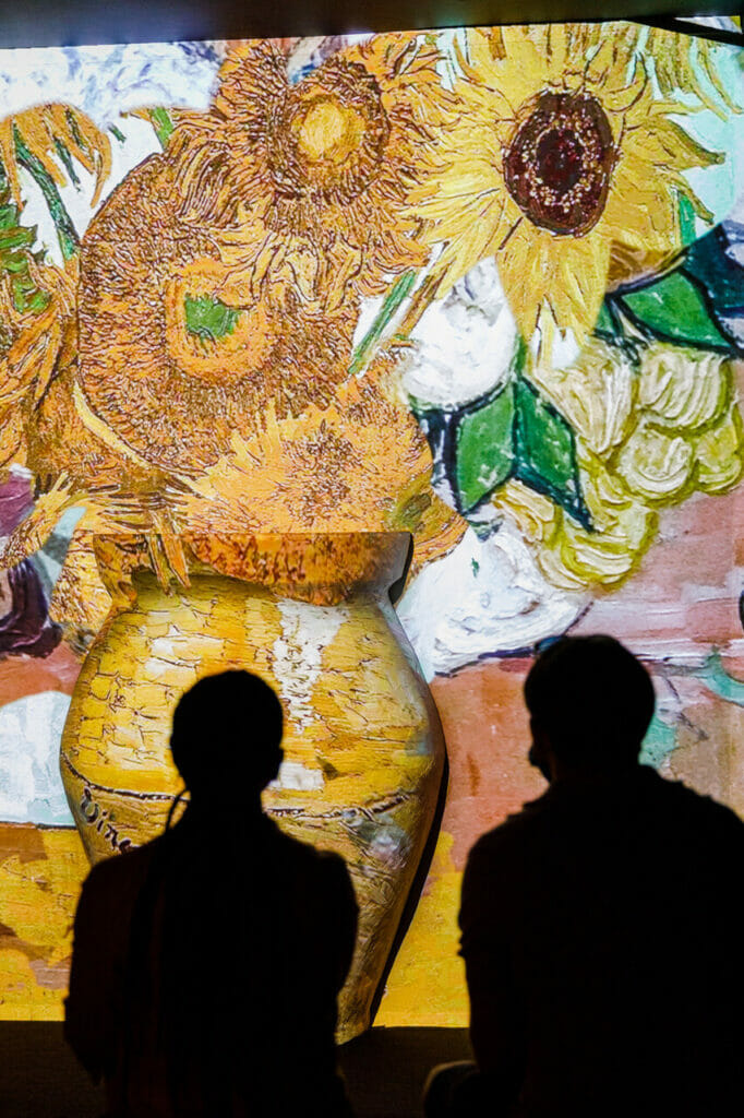Two people admiring the projection on the wall of the famous Vase with 12 sunflowers by Van Gogh at Van Gogh Immersive Experience London