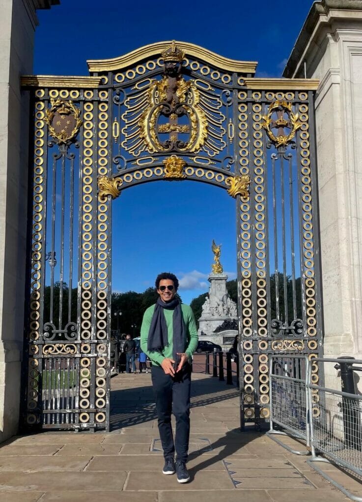 Pericles Rosa wearing sunglasses, a green sweater, blue scarf, marine blue trousers and sneakers passing through a St. James's Park gate, London, and the Queen Victoria Memorial in the background