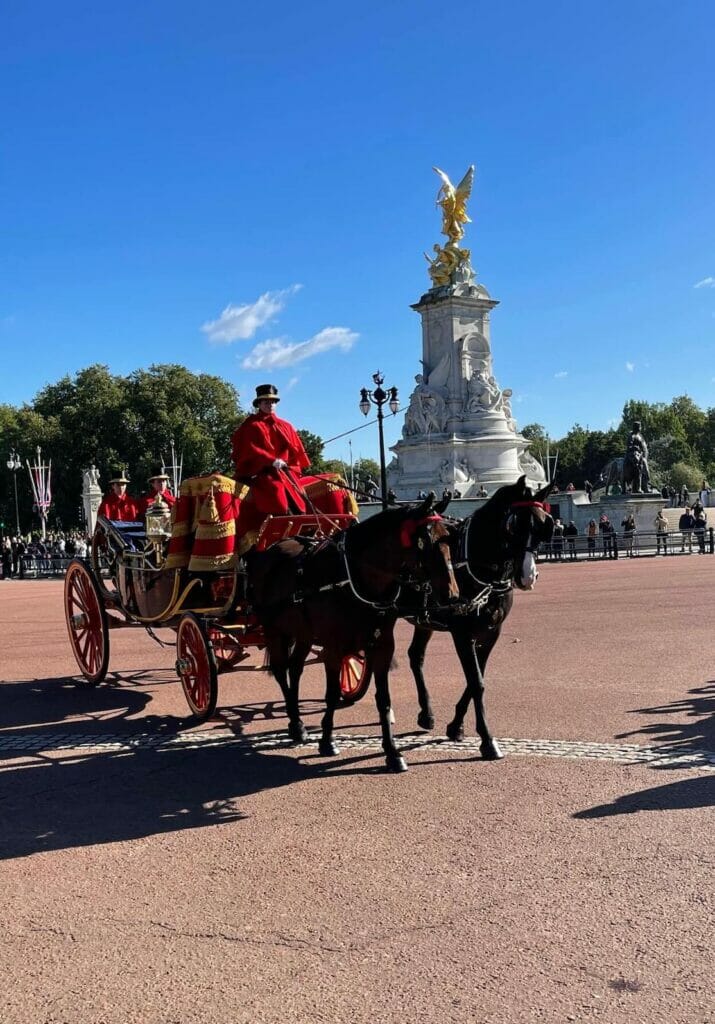 Two horses, a carriage and three men wearing red cape and black hat in front of Queens Victoria Memorial, London, bringing guests to visit Buckingham Palace