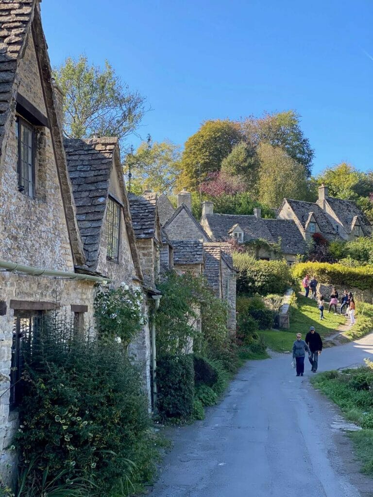 Some people walking on a street bordered by honey-coloured 17th-century stone cottages with steeply pitched roofs in the village of Bibury