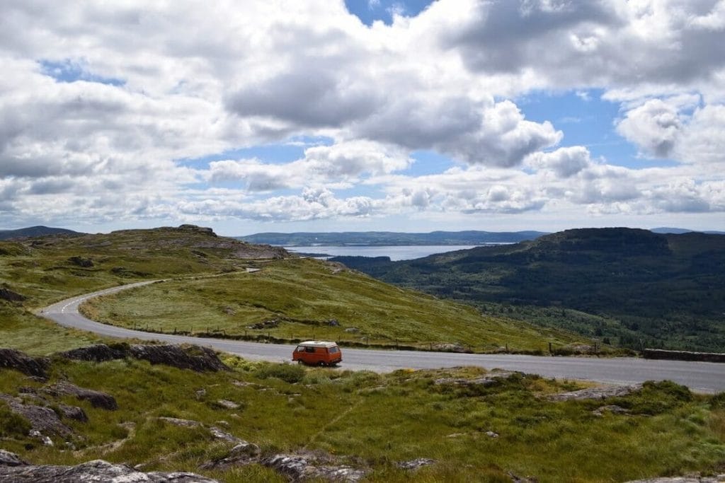 An orange campervan driving on the South West Coastal 300 surrounded by greeny mountainous landscape and a lake in the background.